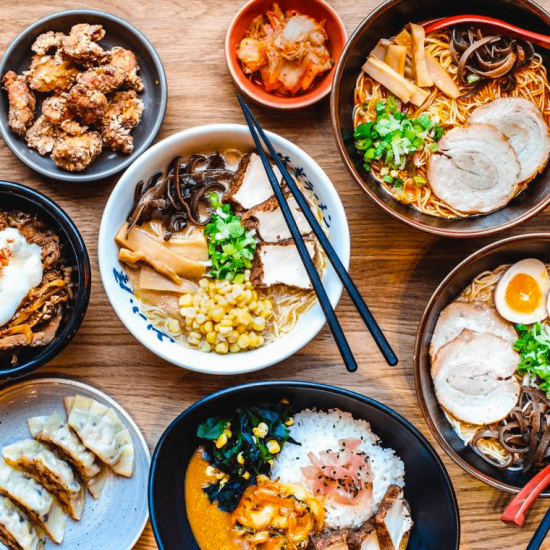 View from above, a wooden table is covered with various bowls and plates, filled with yellow, white and orange vegetables, noodles, and rice