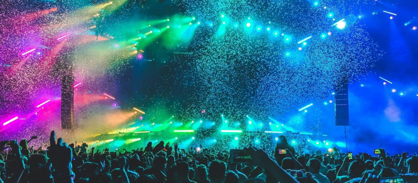 A crowd of people face toward a stage, multi-colored lights and confetti fill the air