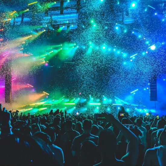 A crowd of people face toward a stage, multi-colored lights and confetti fill the air