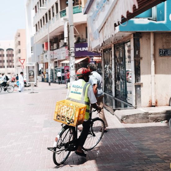 Man rides bicycle with a yellow basket on the back, concrete white buildings in the